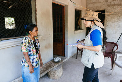A woman in an UNA-UK vest talks to a woman facing her. They are standing in a verandah of a house.