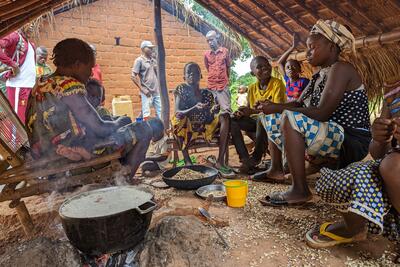 Women and children sit under an informal shelter covered by grass mats in what appears to a cooking area. A pot of rice is cooking on a firewood stove. 