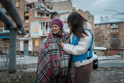 Olena, 85, stands outside her damaged home in Kharkiv, Ukraine, supported by humanitarian aid after a May 2024 attack.